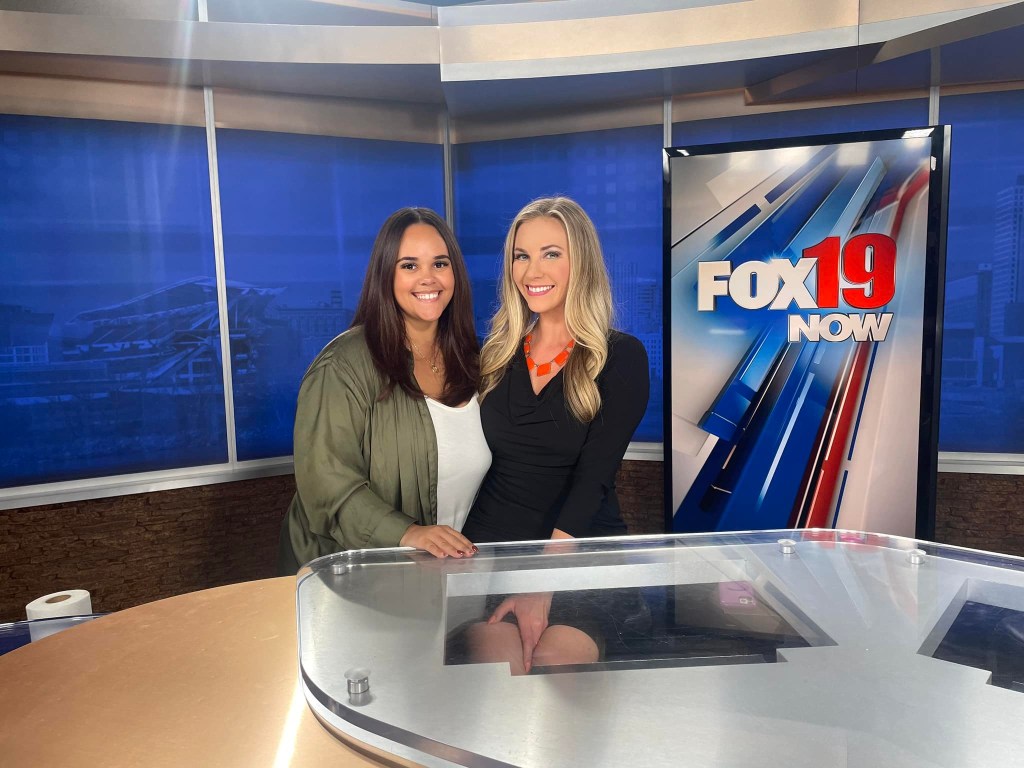 Two women in front of a news desk, with a blue wall and a sign that says "FOX19 NOW" behind them.