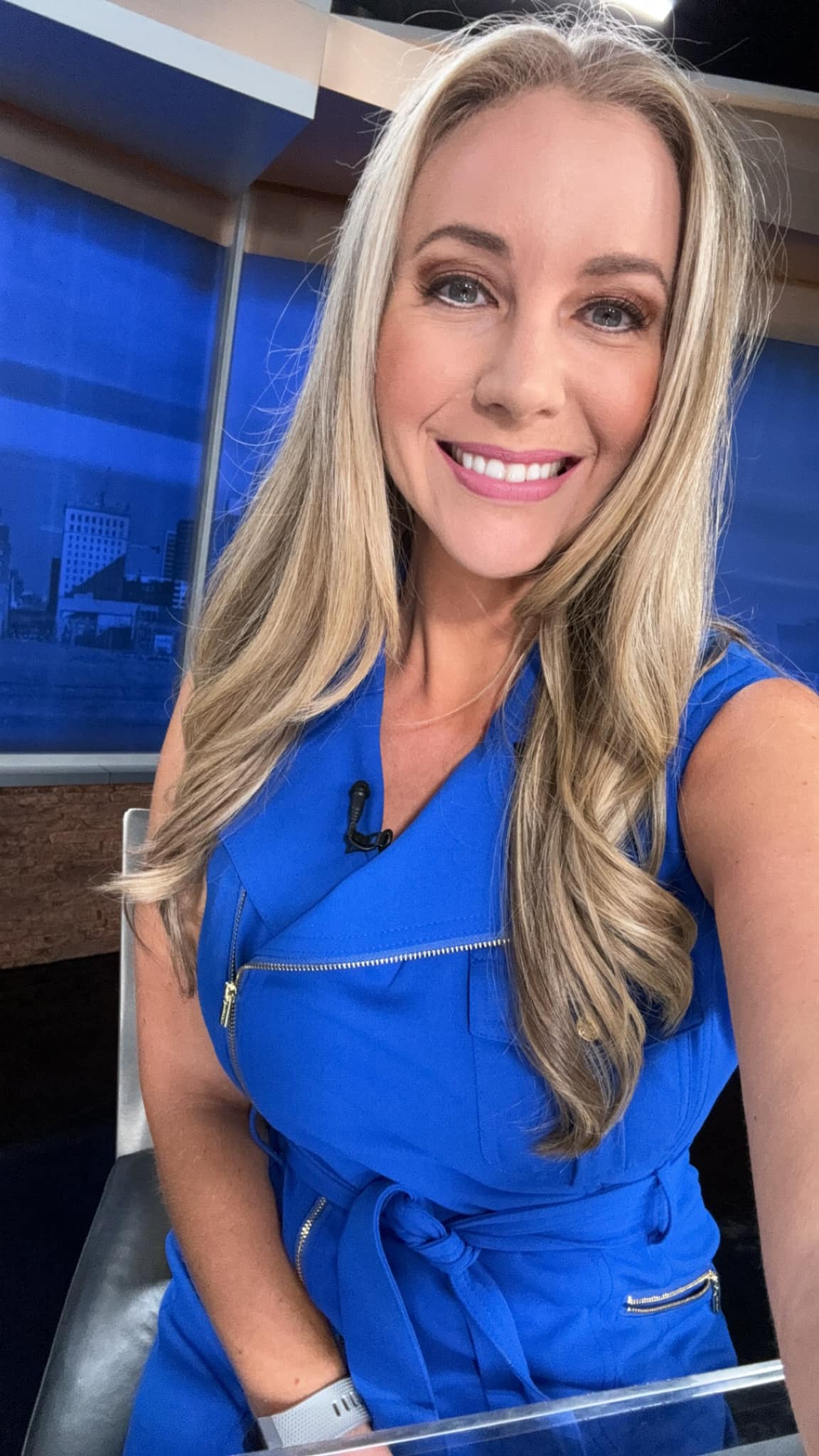 A woman with long blonde hair and a blue dress is sitting in a chair, possibly in a news studio.