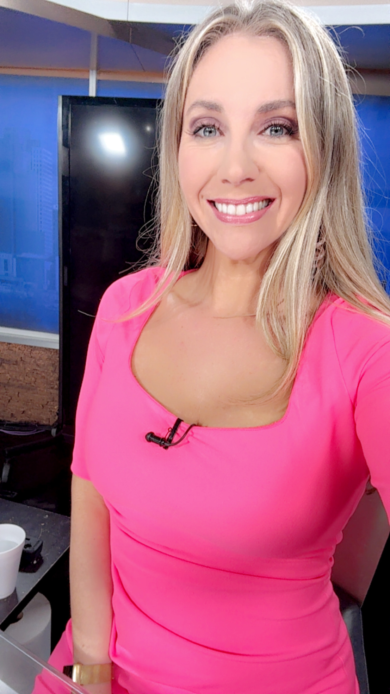 A blonde woman in a pink dress is taking a selfie in a newsroom or studio setting. She is standing in front of a black TV ...