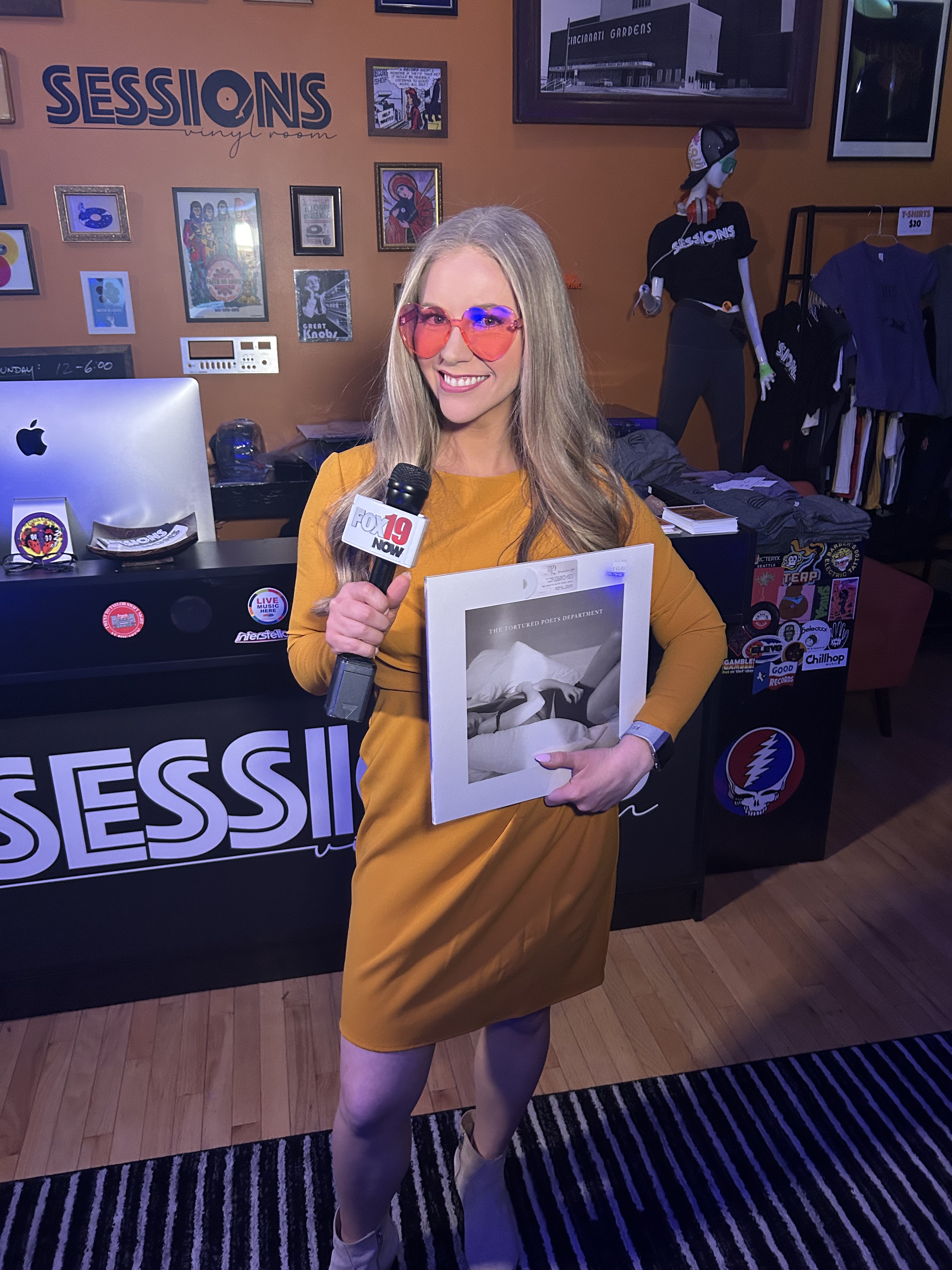 A woman in a yellow dress holds a microphone and album, standing in front of a record store counter with various items on ...